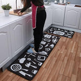 Bas du corps de dame en pantalon noir et tablier rose debout sur un tapis de cuisine noir et blanc aux motifs ustensiles de cuisine, avec un identique plus petit à coté, dans une cuisine blanche