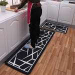 Jambes de dame en pantallon noir debout su tapis de cuisine noir et blanc aux motifs géometriques, un tapis identique à coté plus petit,dans une cuisine blanche